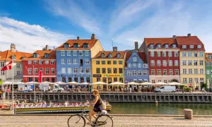 En kvinna cyklar längs Nyhavn i Köpenhamn, ett soligt exempel på danmark klimat. Färgglada hus och kanalbåtar skapar en livlig atmosfär.