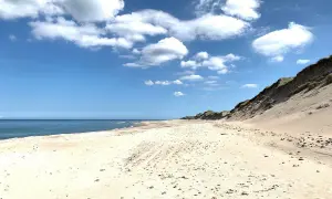 Sandy beach with dunes, calm blue sea, and white clouds under a bright blue sky.