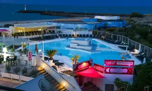 Kvällsbild över en poolklubb vid Cap d'Agde. Ljusblå pool med solstolar, palmer och en scen i mitten. Havet och en fyr syns i bakgrunden.