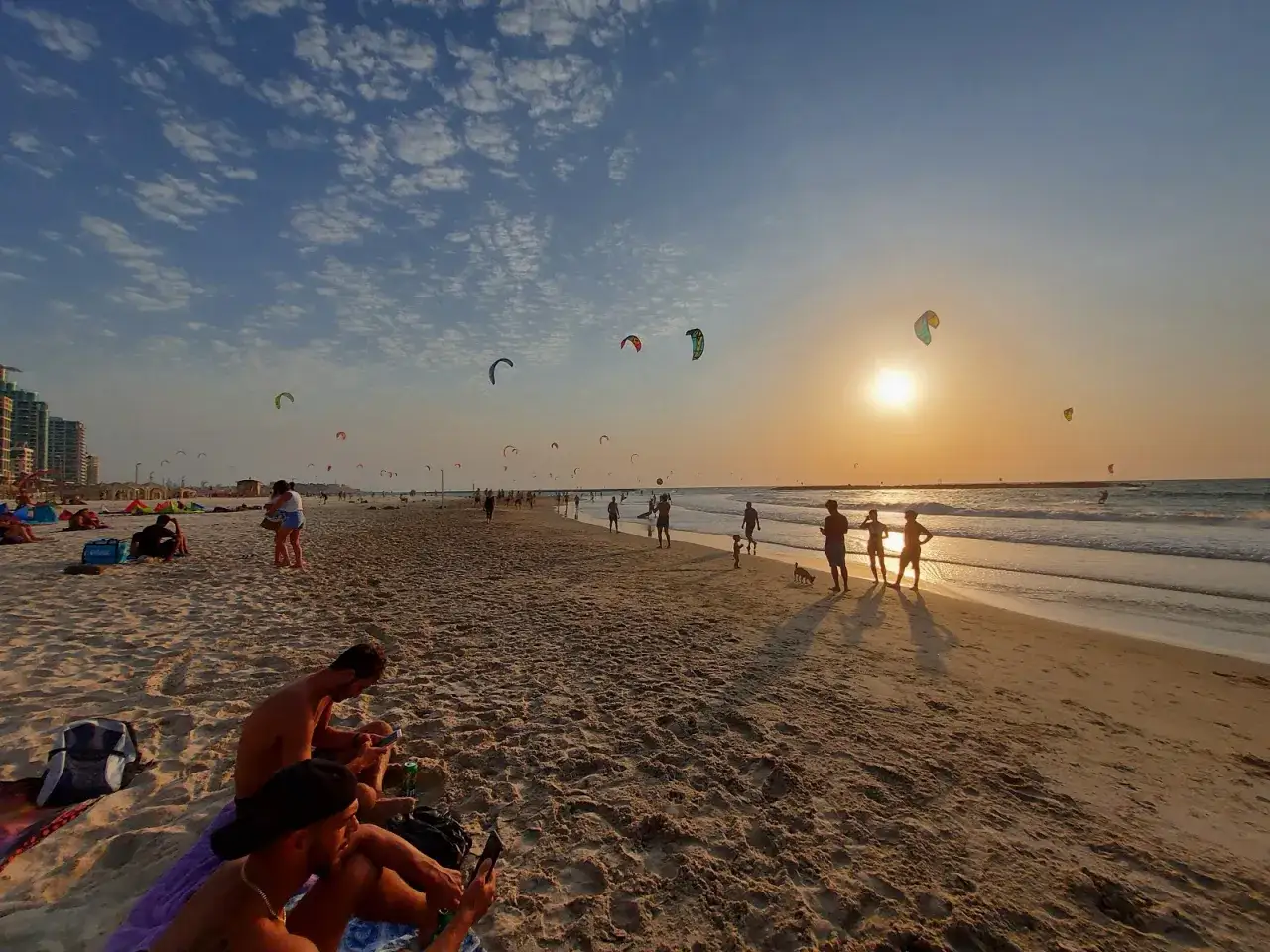Solnedgång över stranden i Tel Aviv med många kitesurfare i luften. Människor njuter av kvällen på sanden.