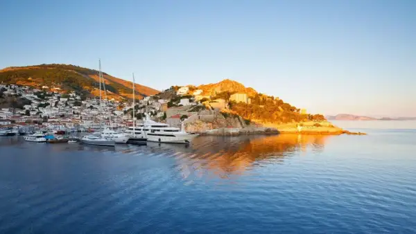 Solnedgång över hamnen på Hydra, Grekland. Lyxyachter guppar i det blå vattnet framför en vit stad som klättrar uppför en grönskande kulle.