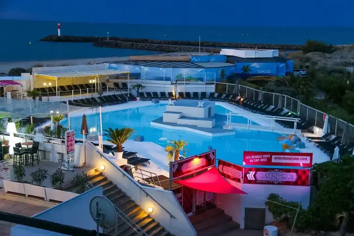 Kvällsbild över en poolklubb vid Cap d'Agde. Ljusblå pool med solstolar, palmer och en scen i mitten. Havet och en fyr syns i bakgrunden.