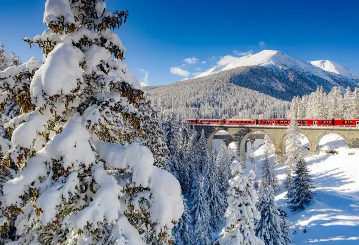 Röda tåget korsar en snöig bro i Schweiz, ett drömresmål med majestätiska berg och vinterlandskap.